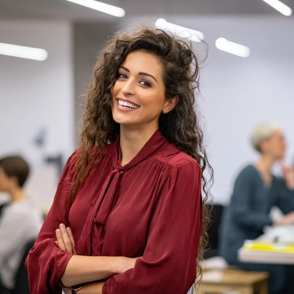 Successful businesswoman standing in creative office and looking at camera. Young latin woman entrepreneur in a coworking space smiling. Portrait of beautiful business woman standing in front of business team at modern agency with copy space.